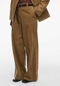 Bruine corduroybroek met een wijde pasvorm, voorzien van verticale ribbels, riemlussen en voorzakken. Gepaard met zwarte schoenen.