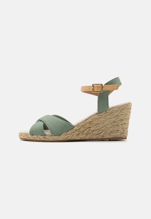 Green fabric wedge sandal with crisscross straps, tan ankle strap with a buckle, and a textured jute-wrapped heel.