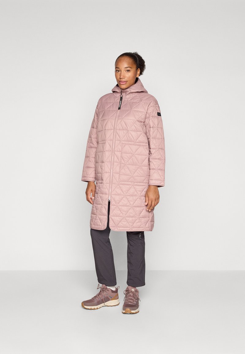 Manteau matelassé rose clair avec une capuche, doté d'une fermeture éclair sur le devant, de manches longues et d'une coupe mi-longue, associé à un pantalon sombre et à des chaussures de randonnée marron.