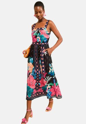 Frau trägt ein buntes Midikleid mit Blumenmuster und breiten Trägern, hält eine gewebte Clutch und trägt rosa Blumen-Sandalen und Ohrringe.