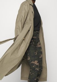 Khaki trenchcoat med en avslappnad passform, knappdetaljer, i kombination med tajta kamouflerade byxor i gröna och bruna nyanser.