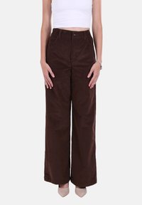 Pantaloni marroni in velluto a coste a gamba larga con vita alta, tasche laterali e chiusura con bottone metallico. Texture liscia con dettagli sottili a coste.