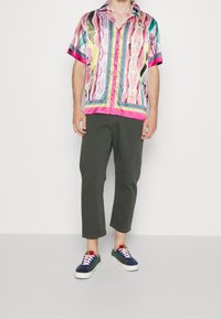 Mann in einem kurzärmeligen, mehrfarbigen, gemusterten Hemd, dunklen, knöchellangen Hosen und marineblauen Sneakers mit weißen Sohle und Schnürsenkeln, der auf einem grauen Boden steht.