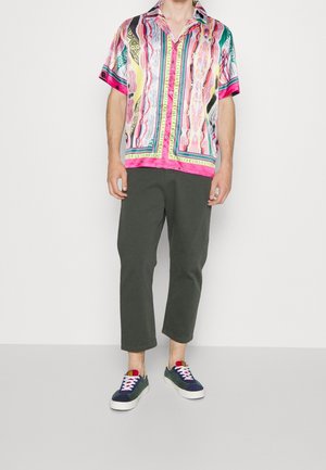 Uomo che indossa una camicia a maniche corte a fantasia multicolore, pantaloni scuri a maniche corte e sneaker blu navy con suole e lacci bianchi mentre sta in piedi su un pavimento grigio.