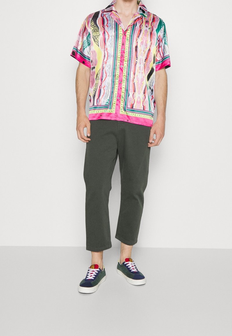 Mann in einem kurzärmeligen, mehrfarbigen, gemusterten Hemd, dunklen, knöchellangen Hosen und marineblauen Sneakers mit weißen Sohle und Schnürsenkeln, der auf einem grauen Boden steht.
