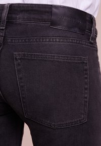 Černé džíny vyrobené z denimu se slim fit střih a šitými detaily, s koženou nášivkou na zadním pásku. Viditelné jsou dvě zadní kapsy.