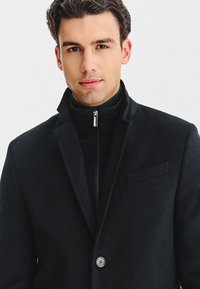 Czarny wełniany płaszcz o dopasowanym kroju, z zapięciem na jeden guzik i klapami w szpic, noszony na czarnym swetrze z wysokim kołnierzem.