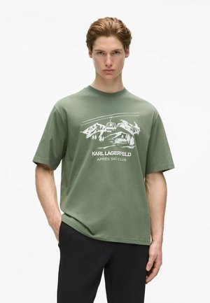Junger Mann mit hellbraunen Haaren trägt ein grünes Karl Lagerfeld T-Shirt mit verschneiten Bergen, einem Skilift und dem Text "Après Ski Club."