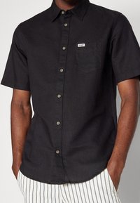 Chemise noire à manches courtes avec boutons, confectionnée dans un tissu léger, dotée d'une poche poitrine et d'une petite étiquette de marque. Finition texturée.