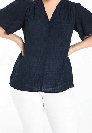 Woman wearing a dark textured short-sleeve blouse with a V-neck, paired with white pants, hands resting on hips.