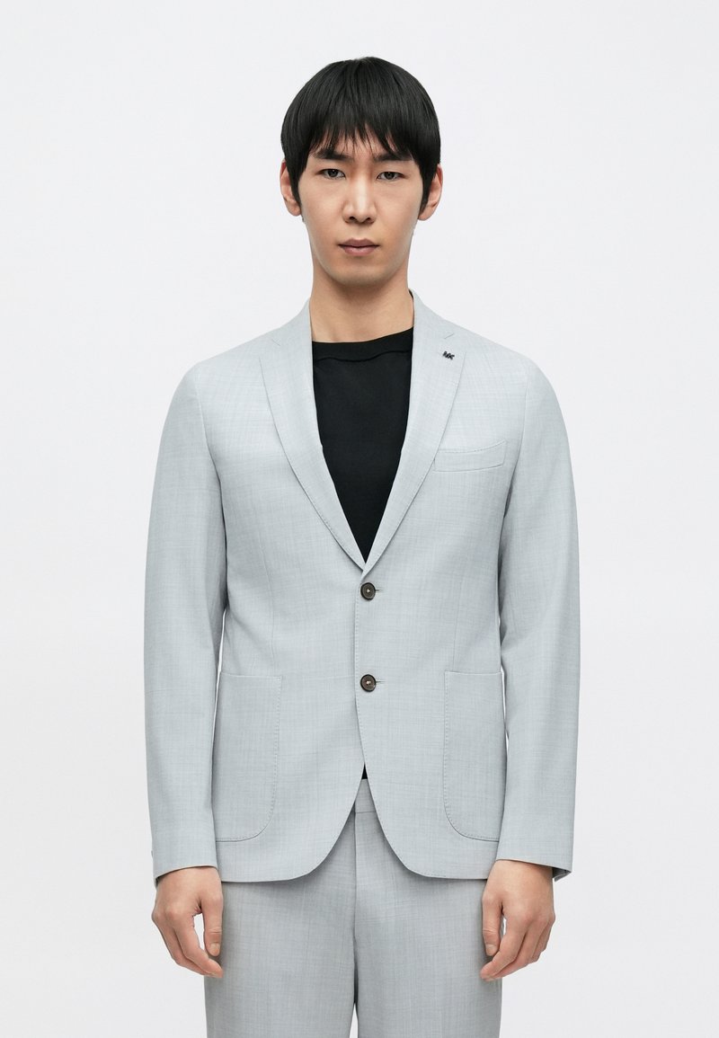 Világosszürke öltönyzakó bevágott hajtókával, két első zsebbel és barna gombokkal. Fekete, texturált felső rész fölött viselve.