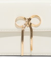 Crème kleurige lakleren tas met een gouden metallic strikaccent en lange lintpunten. De gladde textuur en strakke lijnen versterken het ontwerp.