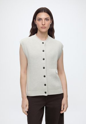 Femme aux longs cheveux foncés portant un gilet en tricot beige sans manches boutonné et un pantalon sombre, debout devant un fond uni.