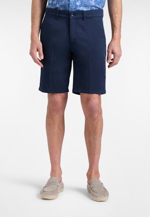 Uomo che indossa pantaloncini blu navy fino al ginocchio, scarpe beige slip-on e una camicia floreale blu, in piedi su uno sfondo bianco.