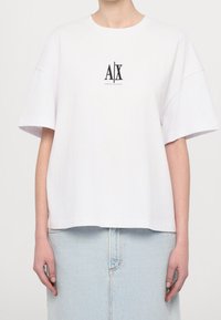 T-shirt blanc oversize avec logo noir "A|X Armani Exchange" sur la poitrine, porté par une personne portant une jupe en jean bleu clair.