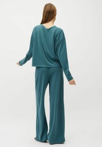 Vrouw die met de rug naar de camera staat, gekleed in een teal kleurige lange mouwen top en wijde broek, op een effen witte achtergrond.