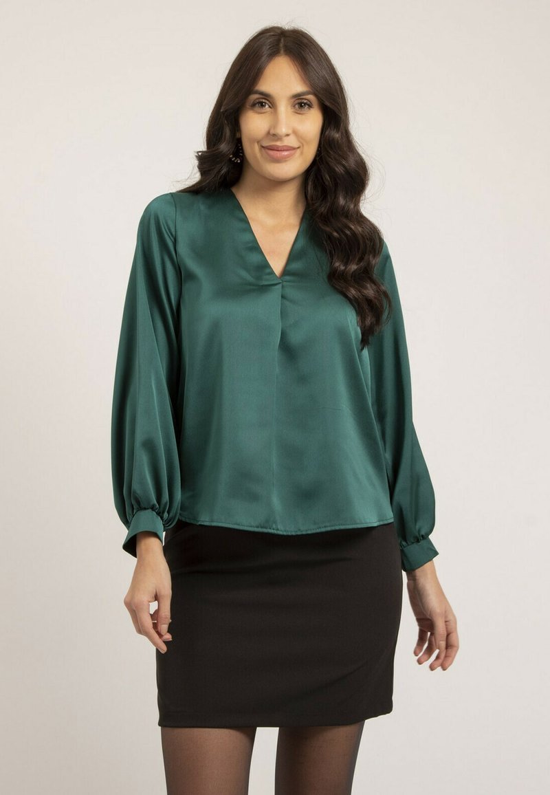 Blouse en satin teal avec un décolleté en V, des manches bouffantes et des poignets élastiques, associée à une jupe noire ajustée et des collants transparents.