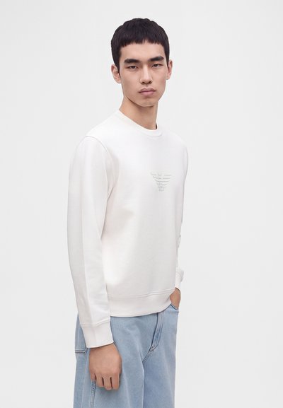 Jeune homme portant un sweat-shirt blanc à manches longues avec un logo discret sur la poitrine et un jean bleu clair, debout devant un fond blanc uni.