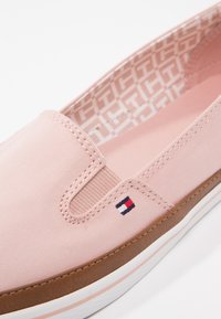 Zapato rosa claro sin cordones con una parte superior de tela texturizada, suela marrón contrastante y un pequeño logo de marca en rojo, blanco y azul en el lateral.