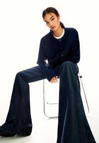 Marineblaue Strickjacke mit rundem Halsausschnitt und Knöpfen, kombiniert mit weit geschnittenen, dunklen Jeans, sitzt auf einem Stuhl aus Metall vor einem hellen Hintergrund.