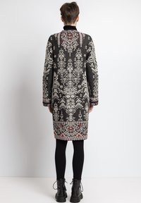 Sort, mønstret lang cardigan med hvide og røde detaljer. Har en høj krave og lange ærmer, tekstureret strikket stof og intrikate motiver.