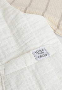 Tela de algodón blanco acolchado con cuadrados cosidos, con una etiqueta que dice "Pequeño y Querido". El fondo muestra una tela texturizada beige clara.