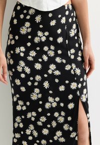 Black midi skirt featuring a daisy floral print in white and yellow. It has a side slit and smooth texture, with a fitted design.