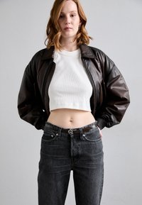 Giacca bomber in pelle marrone, top corto a coste bianco, jeans neri a vita alta con cintura nera lucida, modello che mostra il ventre.