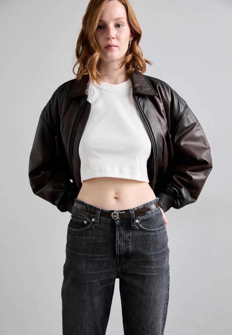 Giacca bomber in pelle marrone, top corto a coste bianco, jeans neri a vita alta con cintura nera lucida, modello che mostra il ventre.