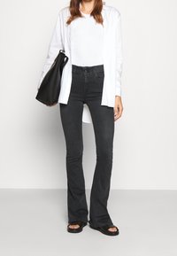 Svarta utsvängda jeans, hög midja med knappstängning, kombinerat med en vit långärmad skjorta och en svart tote-väska. Platta sandaler fulländar looken.