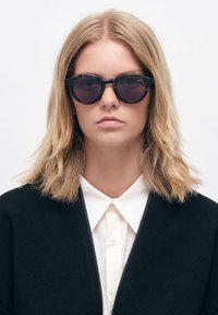 Aurinkolasit mustilla pyöreillä linsseillä, paksulla kehyksellä ja matalla pinnalla. Mallilla, jolla on vaaleanruskeat hiukset ja valkoinen kauluspaita.