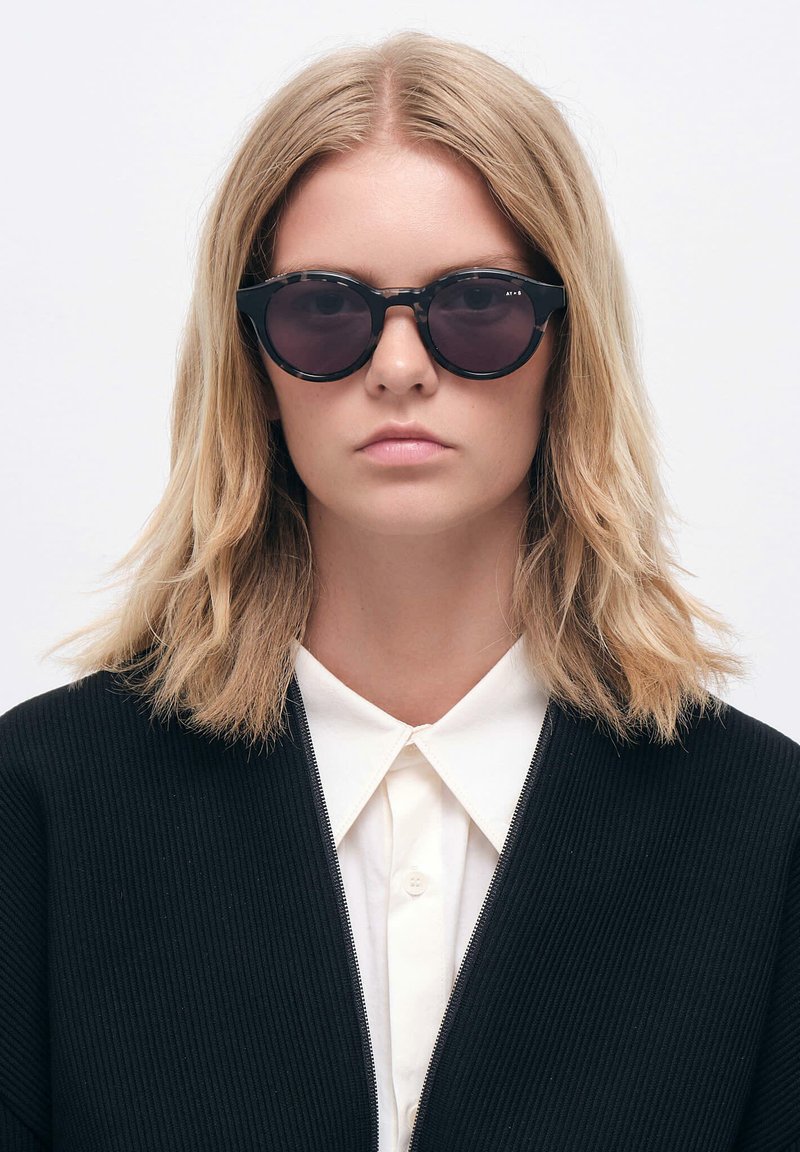 Aurinkolasit mustilla pyöreillä linsseillä, paksulla kehyksellä ja matalla pinnalla. Mallilla, jolla on vaaleanruskeat hiukset ja valkoinen kauluspaita.