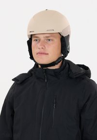 Beiger Skihelm mit Belüftungslöchern und schwarzen Ohrenschützern, getragen von einer Person in einer schwarzen Jacke mit Reißverschlusstasche, vor weißem Hintergrund.