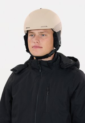 Casque de ski beige avec des trous d'aération et des protège-oreilles noirs, porté par une personne vêtue d'une veste noire avec une poche à fermeture éclair, sur un fond blanc.