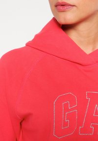 Korálově růžová mikina s měkkou texturou, která má stříbrný detail loga, šňůrky na kapuci a raglánový střih rukávů.