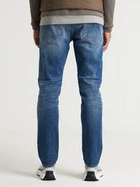 Pantalones vaqueros de mezclilla en azul medio con acentos desvaídos. Presentan un corte recto, diseño de cinco bolsillos y bolsillos traseros cosidos. Zapatillas deportivas grises visibles.