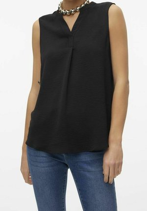 Vrouw draagt een mouwloze zwarte getextureerde top met V-hals en zilveren dikke kralenketting, gecombineerd met blauwe spijkerbroek.