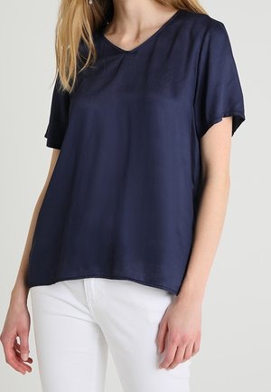 Žena oblečená v volné krátké modré haleně s výstřihem do V v námořnické modři, doplněné bílými kalhotami, zobrazená od ramen po boky.