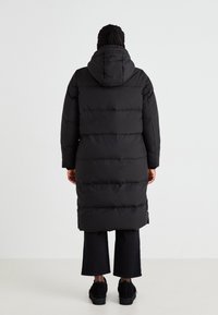 Zwarte pufferjas met een capuchon, voorzien van horizontale doorgestikte secties, lange lengte en zijsplitten. Gedragen over donkere broeken en schoenen.