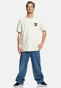 Beige T-Shirt mit kurzen Ärmeln und einem schwarzen DC-Logo auf der Brust, kombiniert mit lockeren blauen Jeans und schwarzen Schuhen.