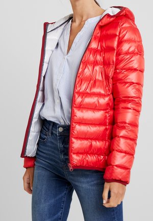 Femme portant une veste matelassée rouge brillante ouverte sur une chemise à rayures bleu clair boutonnée et un jean bleu, debout devant un fond uni.