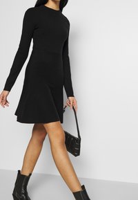 Vestito nero a maniche lunghe con corpino aderente, gonna a campana e texture liscia. Abbinato a una borsa nera e stivaletti ankle.