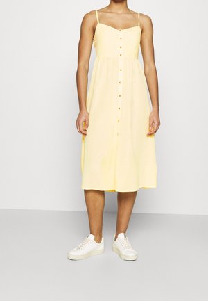 Vrouw draagt een mouwloze gele midi-jurk met knopen vooraan, gecombineerd met witte sneakers, staand tegen een effen lichte achtergrond.