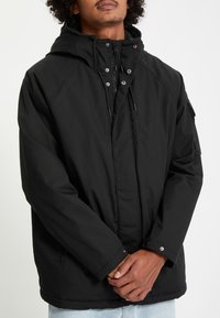 Homme portant une veste d'hiver noire à capuche avec fermeture éclair et boutons-pression, debout les mains jointes à la taille.