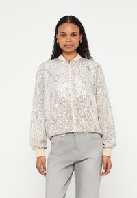 Casaco bomber prateado com lantejoulas, fecho de correr, punhos canelados e gola subida, combinado com jeans cinza claro.