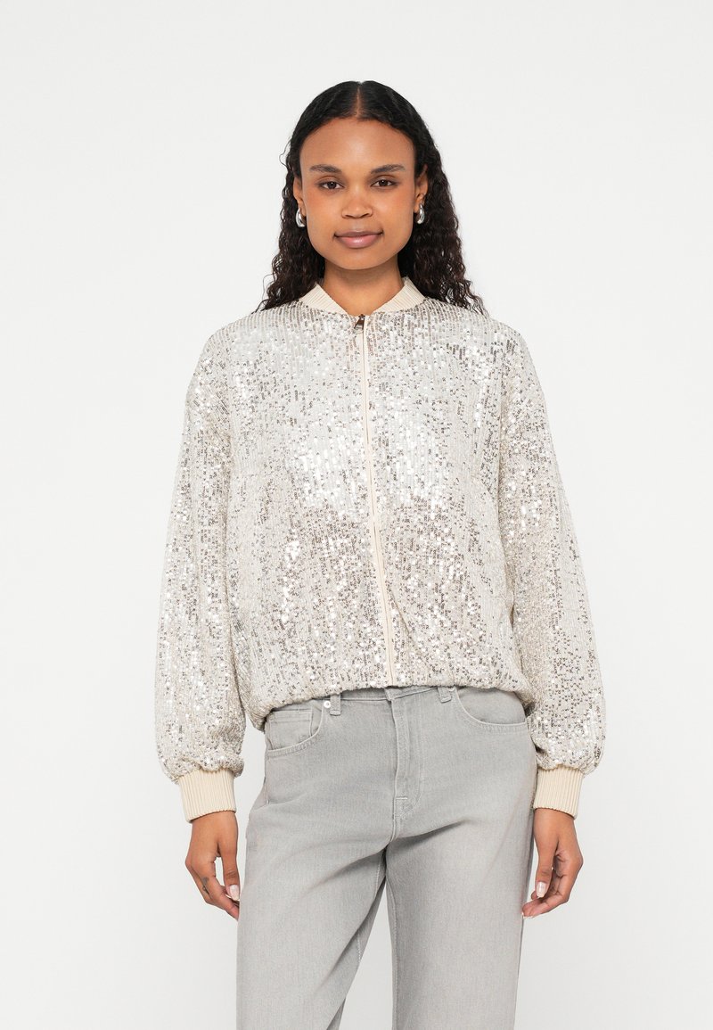 Casaco bomber prateado com lantejoulas, fecho de correr, punhos canelados e gola subida, combinado com jeans cinza claro.
