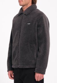 Chaqueta de forro polar gris con cierre de cremallera, cuello y bolsillos laterales. Presenta una superficie texturizada y un pequeño logo en el pecho.