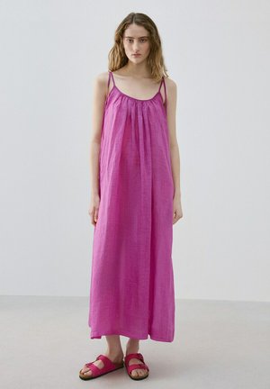 Femme debout vêtue d'une robe ample sans manches magenta à fines bretelles et de sandales assorties à enfiler, posant devant un fond uni.