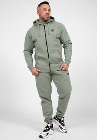 Khaki zip-up hoodie met trekkoorden en voorzakken, gecombineerd met bijpassende joggers met zijzakken en geribde manchetten. Witte en zwarte sneakers.