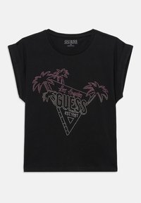 Camiseta de algodón orgánico negra con mangas enrolladas que presenta un gráfico de triángulo de rhinestones, palmeras y el texto "GUESS EST. 1981" en colores vibrantes.
